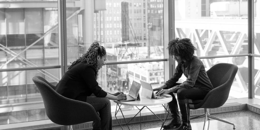 Two women are sat across from one another typing on their laptops. They are sitting in front of a wall of windows and the image is greyscale.