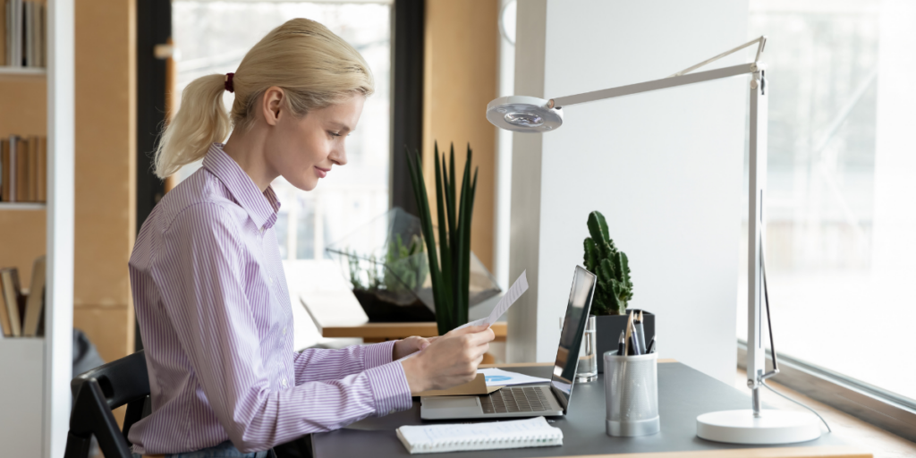 A woman is sat at her laptop and is reviewing a note.