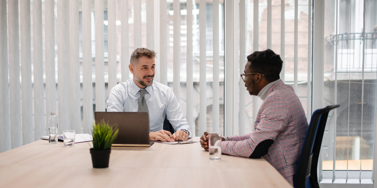 Two men are sat at a boardroom table conversing.
