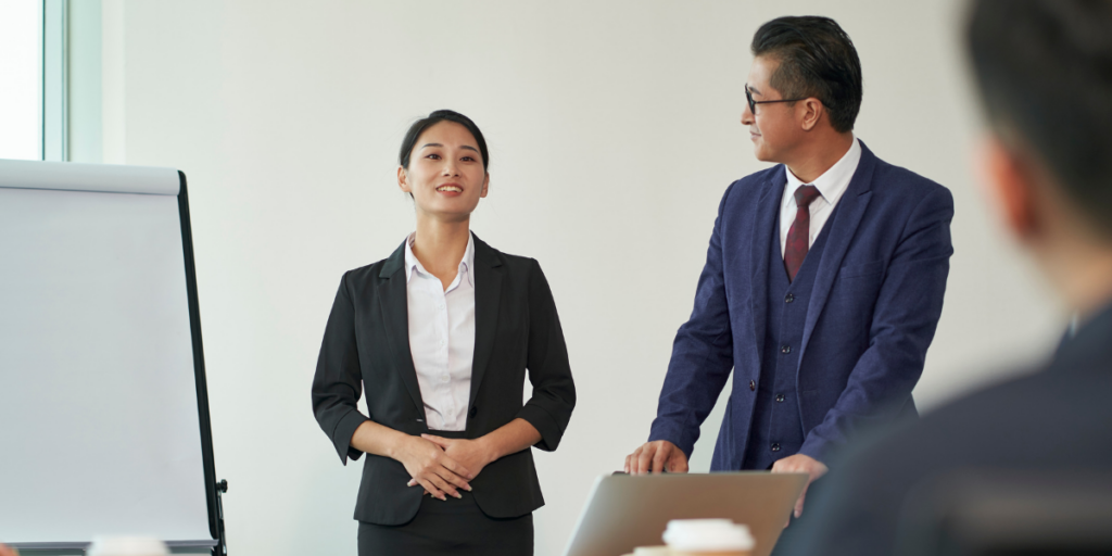 A woman and a man are stood at the front of a boardroom presenting.