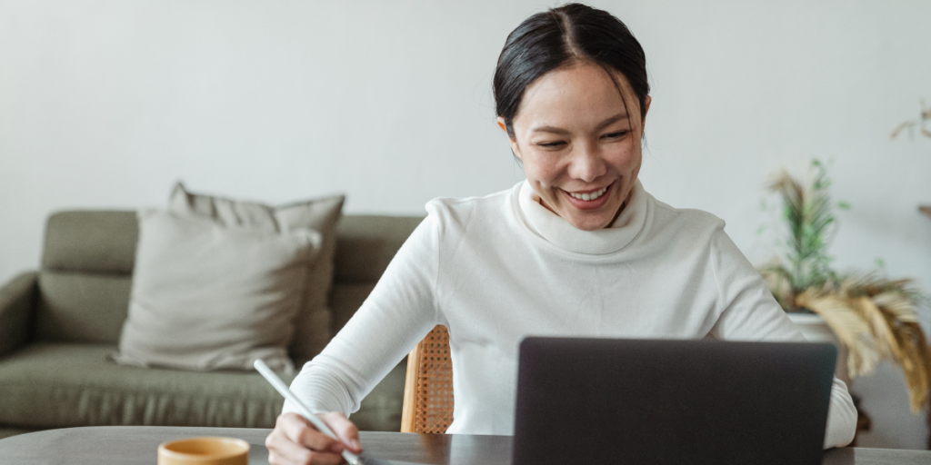 A woman is sat at a table taking notes and working on her laptop.