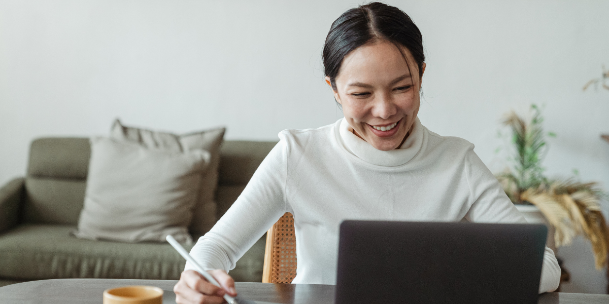 A woman is sat at a table taking notes and working on her laptop.
