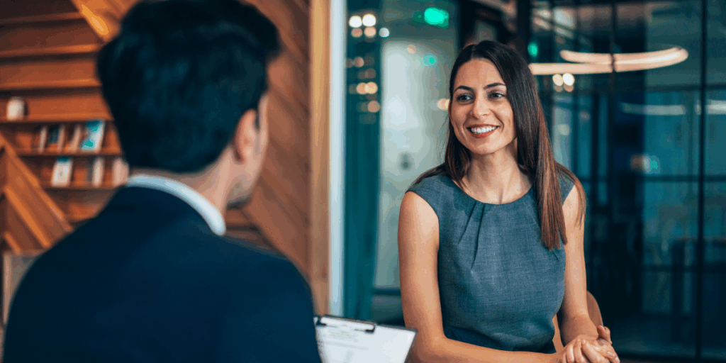 A woman is sat in a boardroom looking across the table at a man holding a clipboard.