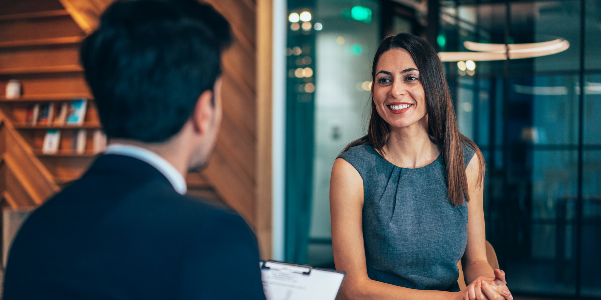 A woman is sat in a boardroom looking across the table at a man holding a clipboard.