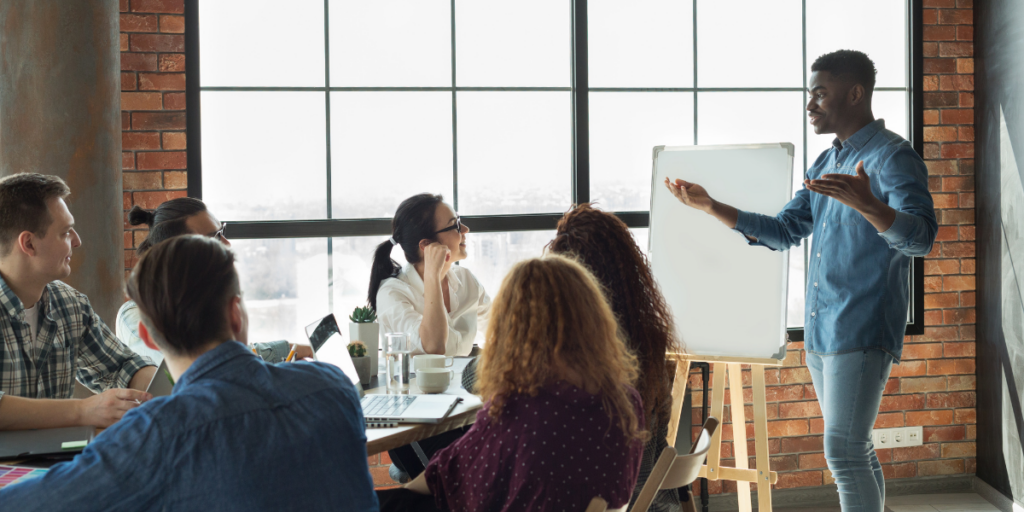A man is standing at the front of a boardroom presenting to a table of colleagues.