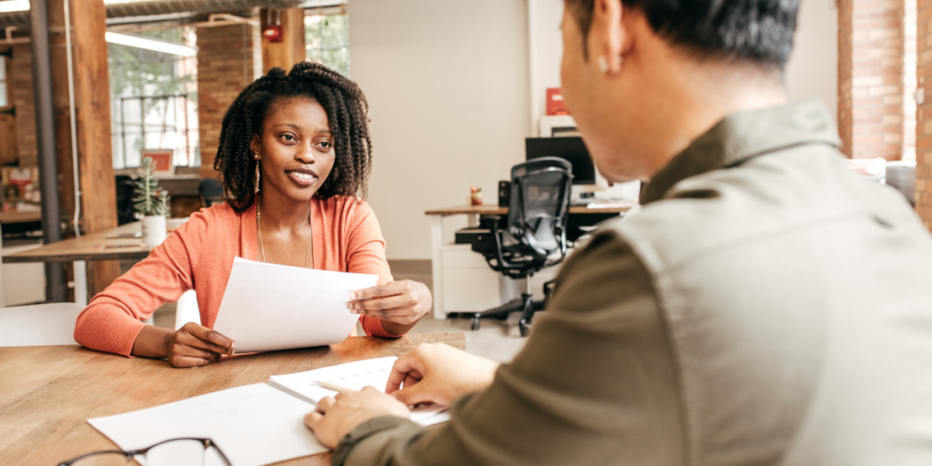 A woman is sat facing a man at a boardroom table and appears to be interviewing him.
