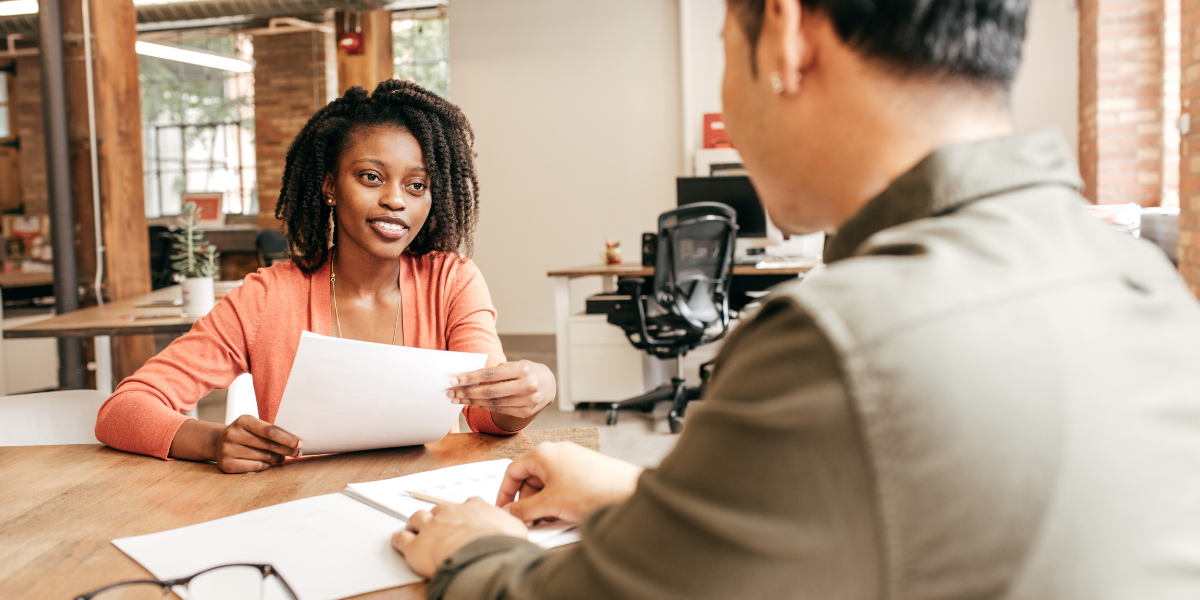 A woman is sat facing a man at a boardroom table and appears to be interviewing him.