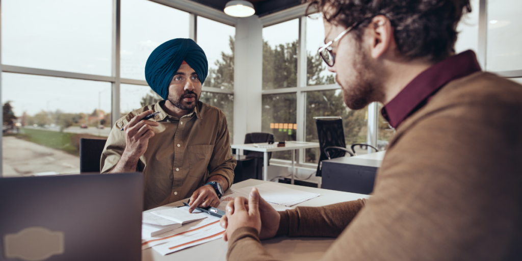 Two men sat across from one another at a desk conversing.
