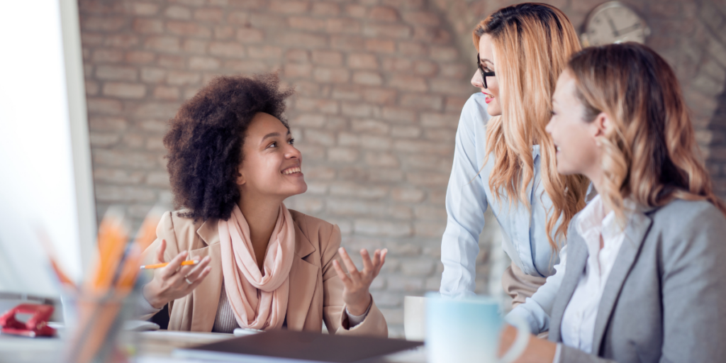 Three people are sat at a desk in an office conversing.