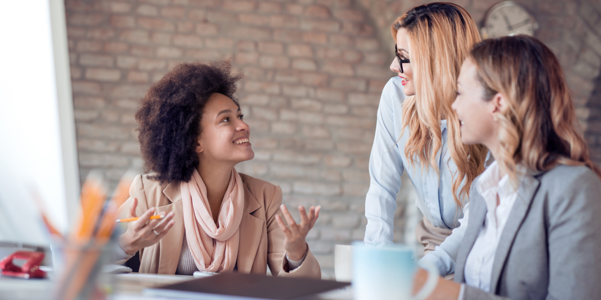 Three people are sat at a desk in an office conversing.