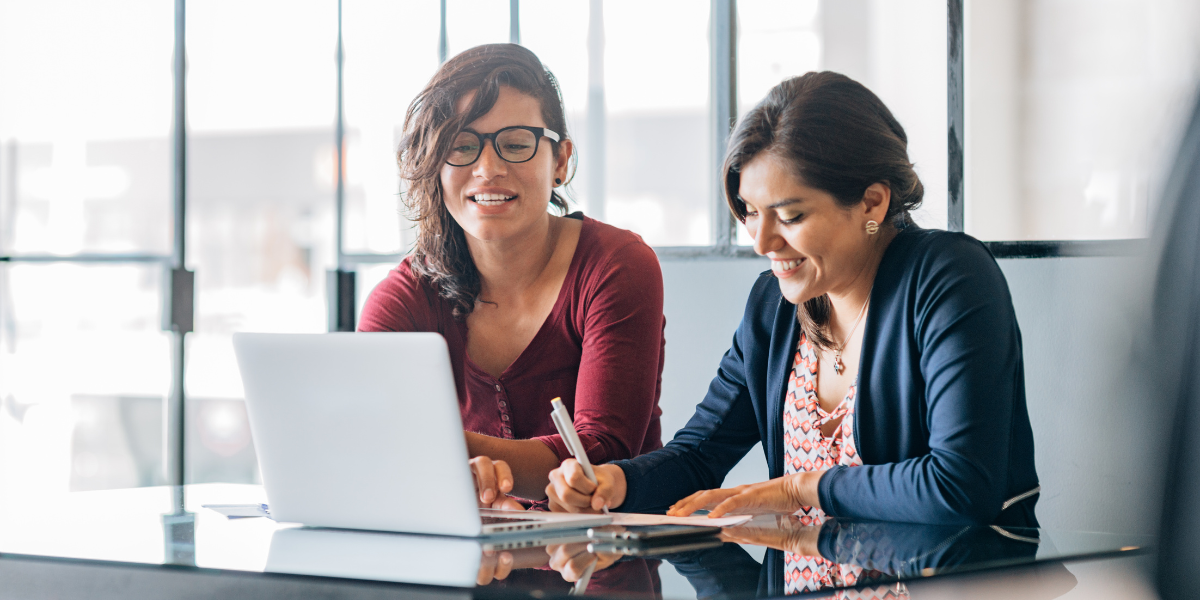 Two women are sat side-by-side conversing in an office space.