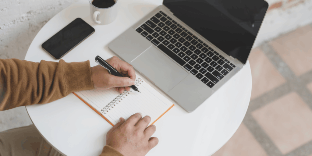 A man is sat at a desk writing notes and looking at a laptop.