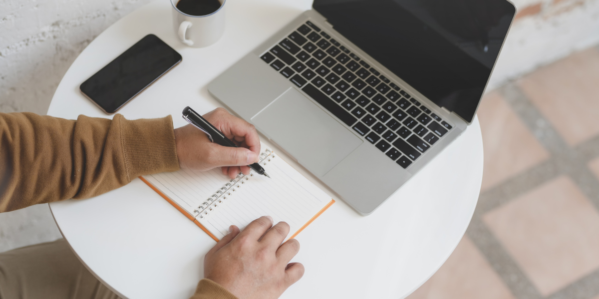 A man is sat at a desk writing notes and looking at a laptop.