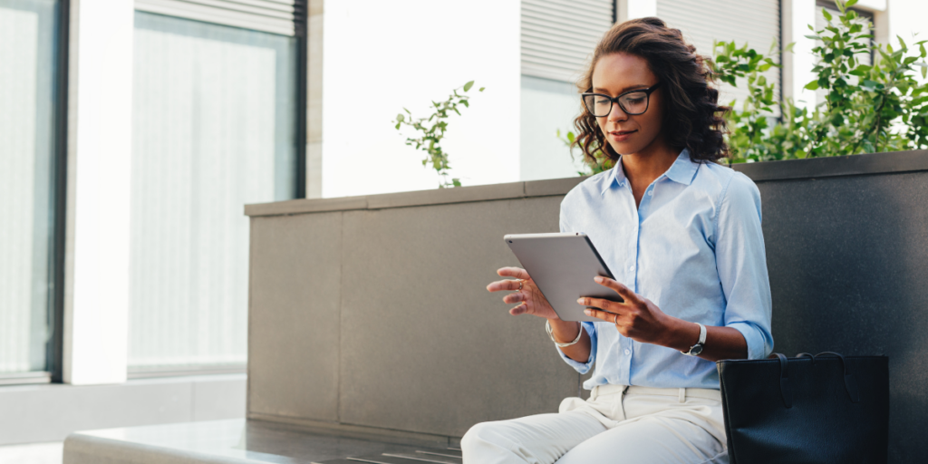 A woman is sat outside on a bench looking at a tablet.