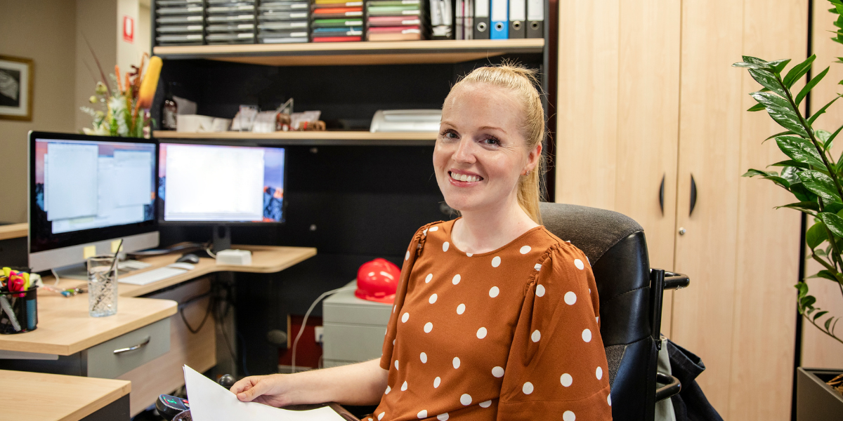 A woman is sat in front of her desk and computer monitor holding papers and smiling at the camera.