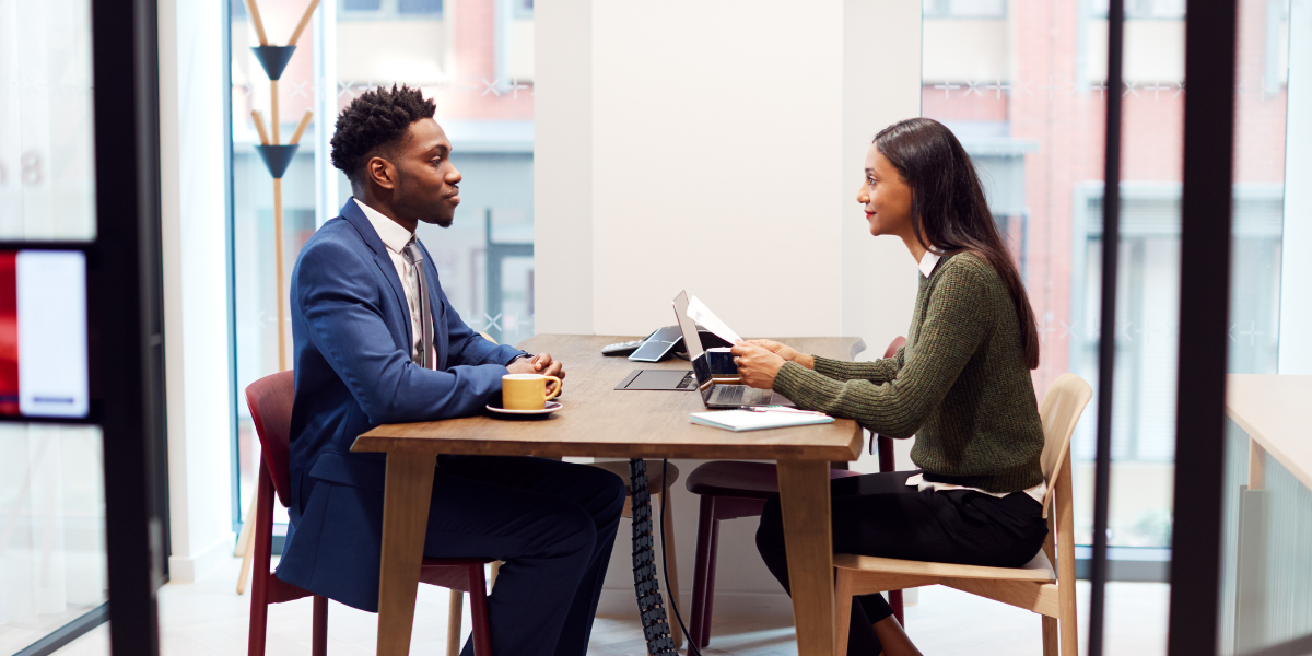 Two people sat across from one another conversing. They are sitting in front of a wall of windows.