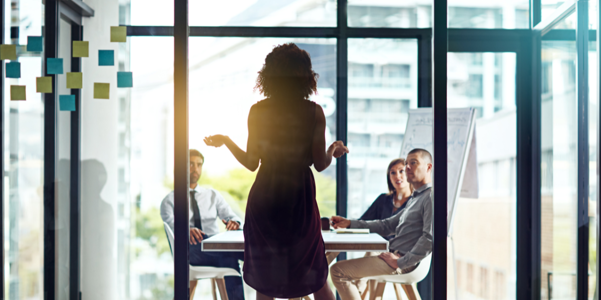 A woman is standing at the head of a boardroom table presenting to a group of colleagues.