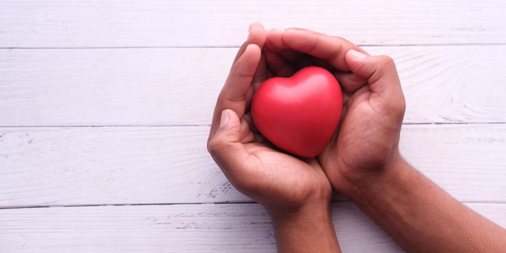 Two hands are holding a foam red heart on a white wooden table.