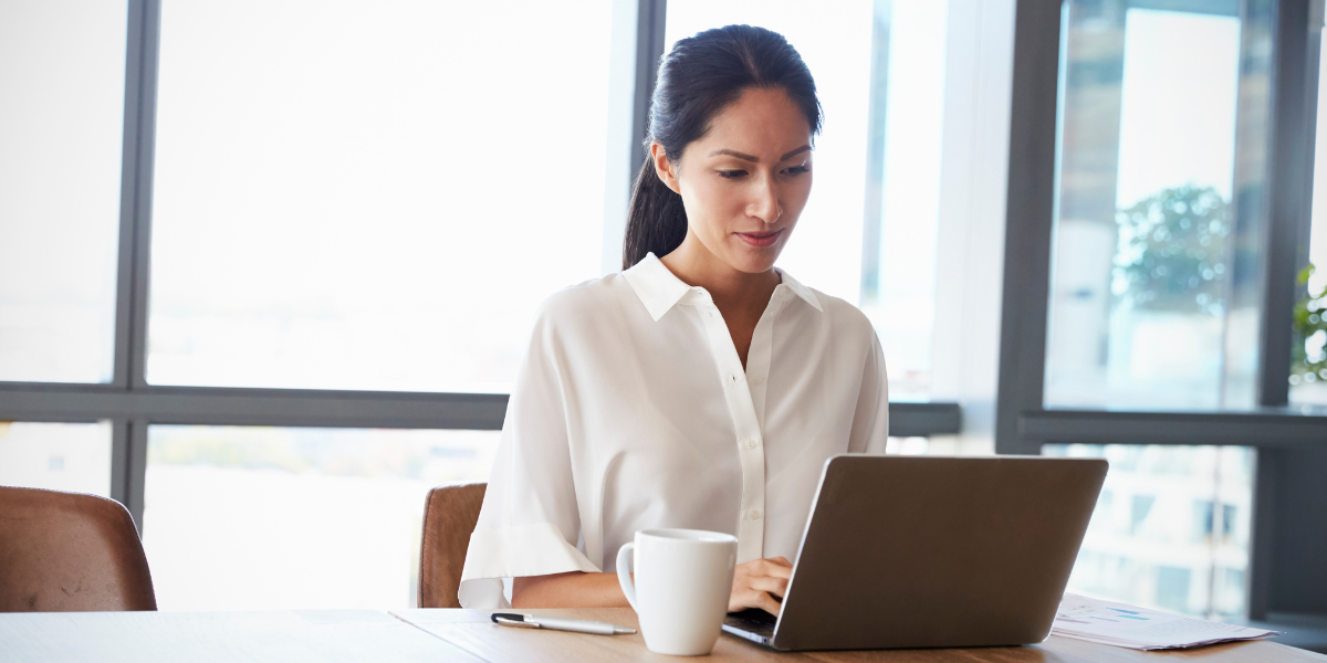 A woman is sitting at a desk typing on her laptop. A wall of windows is behind her.
