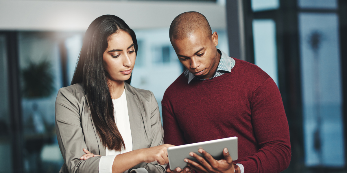 Two people are standing next to one another reviewing a tablet.