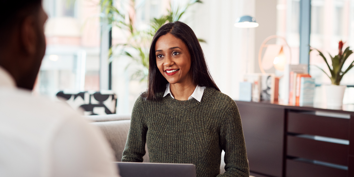 A woman is smiling with a laptop in front of her and she is facing a person across the table.