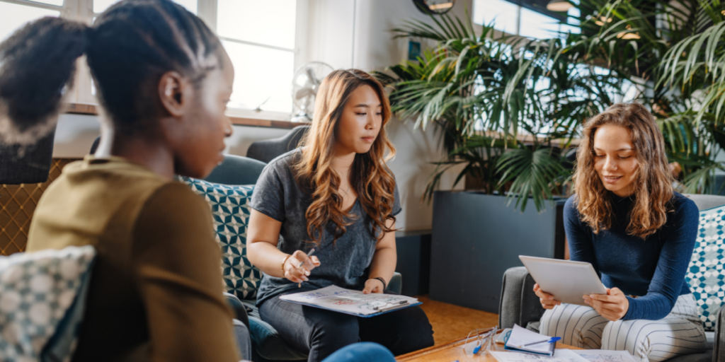 Three women are sat in a circle on chairs reviewing a document and conversing.