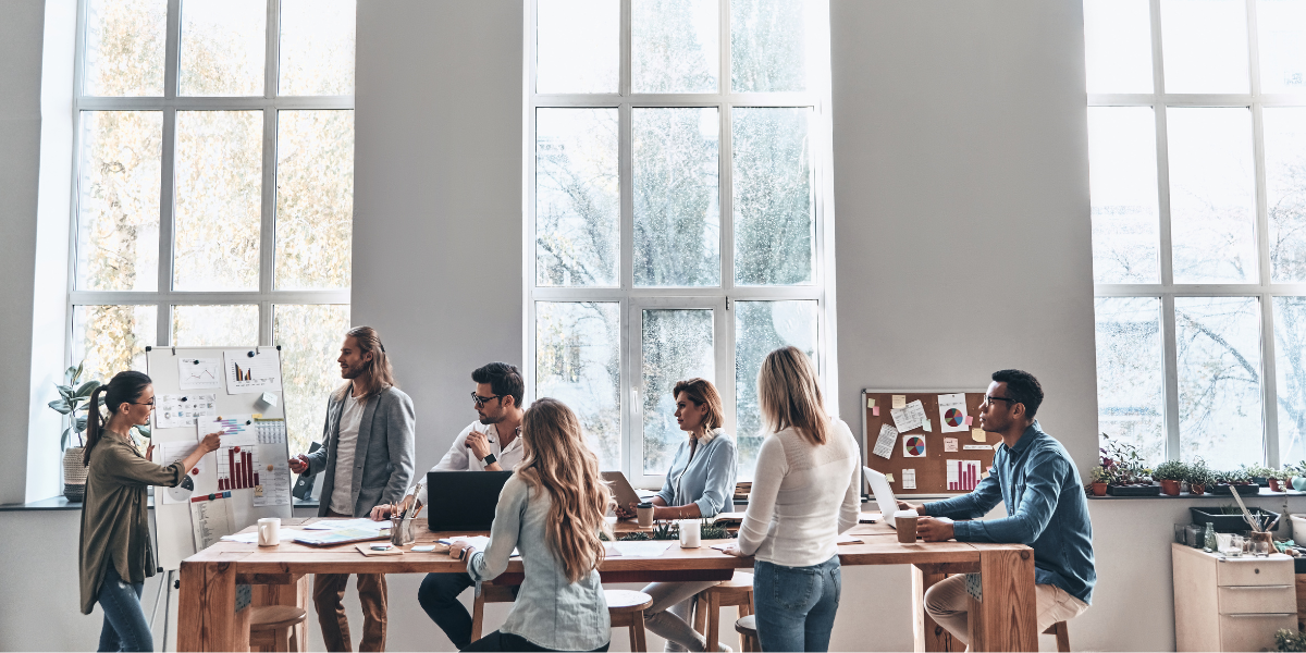 An office with a group of people convened around a table collaborating on a project and conversing.