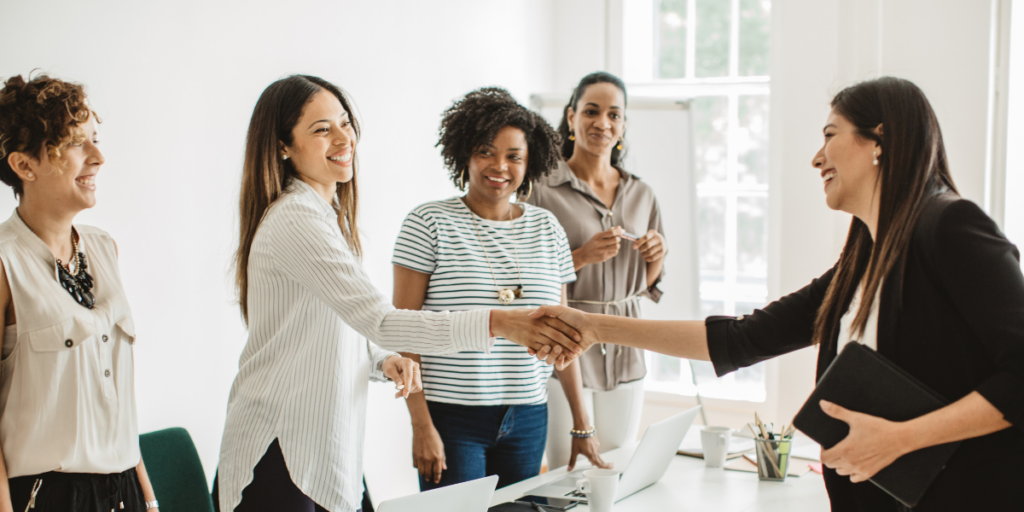 A panel of people are standing on one side of the boardroom table while one woman was being interviewed and is now shaking the hand of the manager.