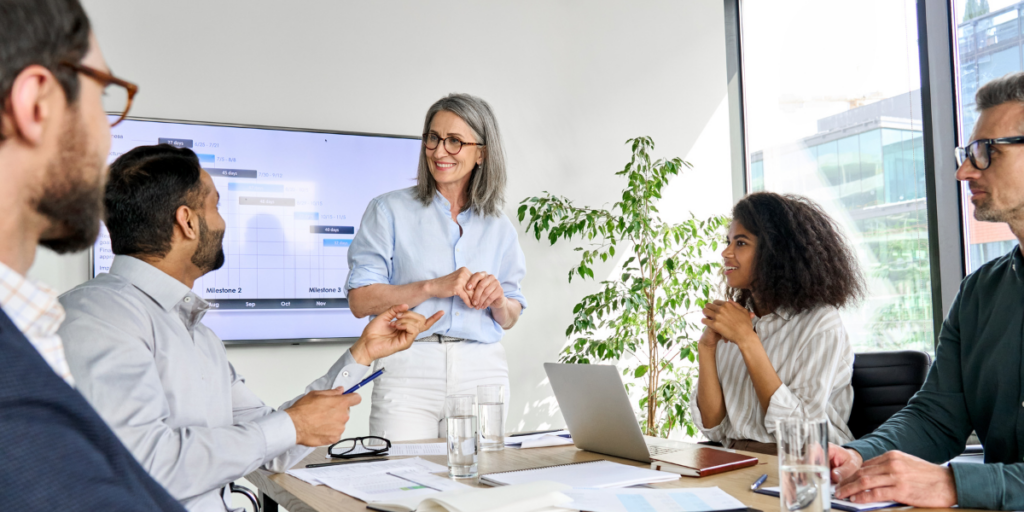 A group of people sitting at a boardroom table while one woman stands at a whiteboard presenting.