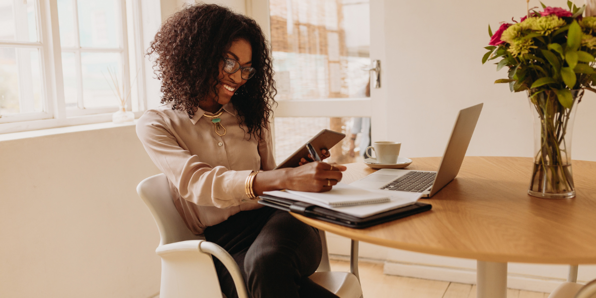 A woman is sat at a desk beside a wall of windows taking notes and looking at her laptop.