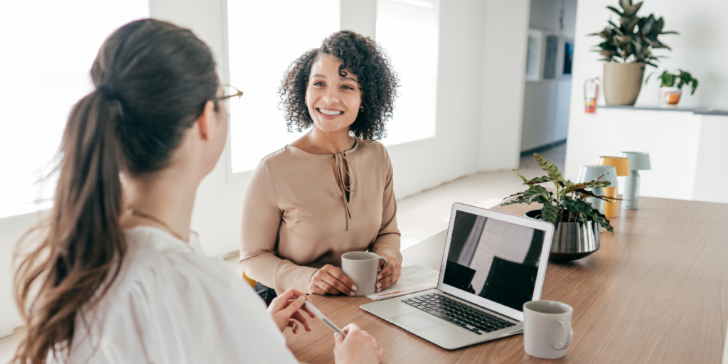 Two women are sat at a boardroom table facing each other conversing.