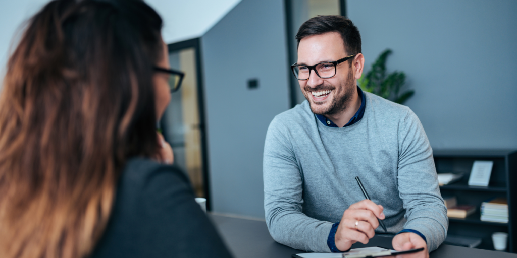 Two people are sat across from each other at a boardroom table conversing.