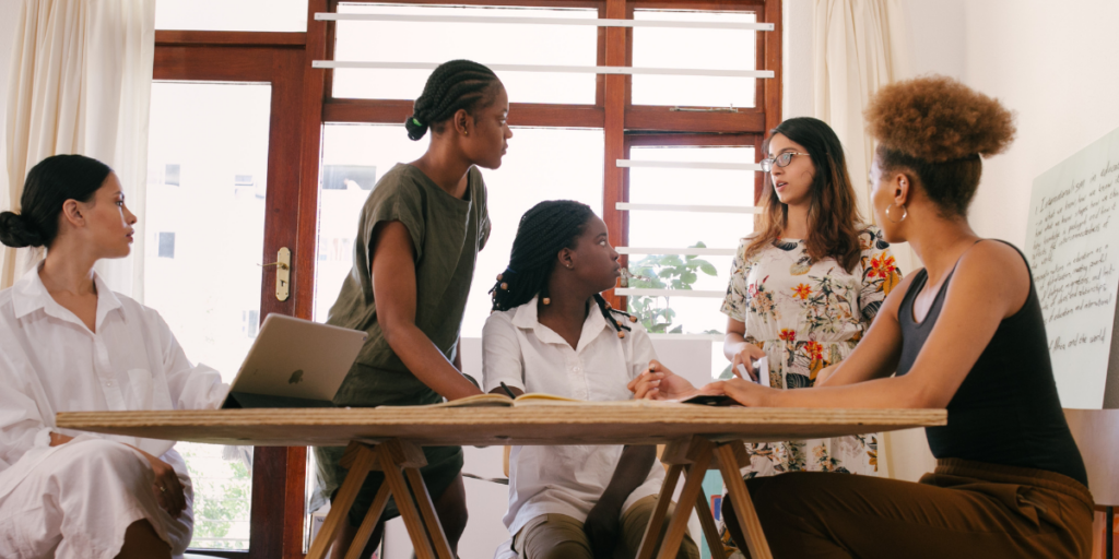 A group of women sitting at a boardroom table while one woman stands at a chart presenting.
