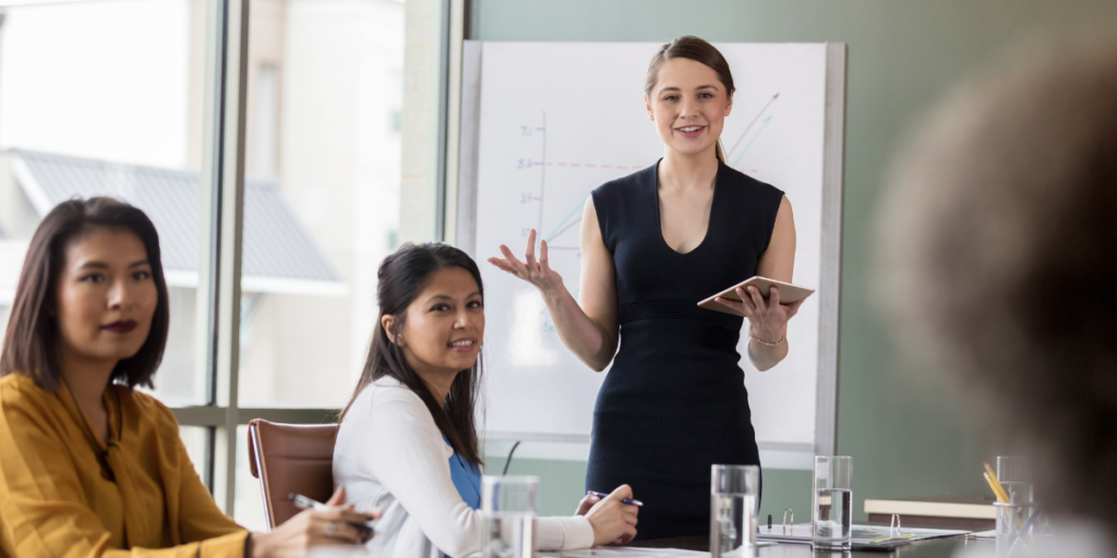 A woman standing holding a tablet and presenting to a boardroom of people.