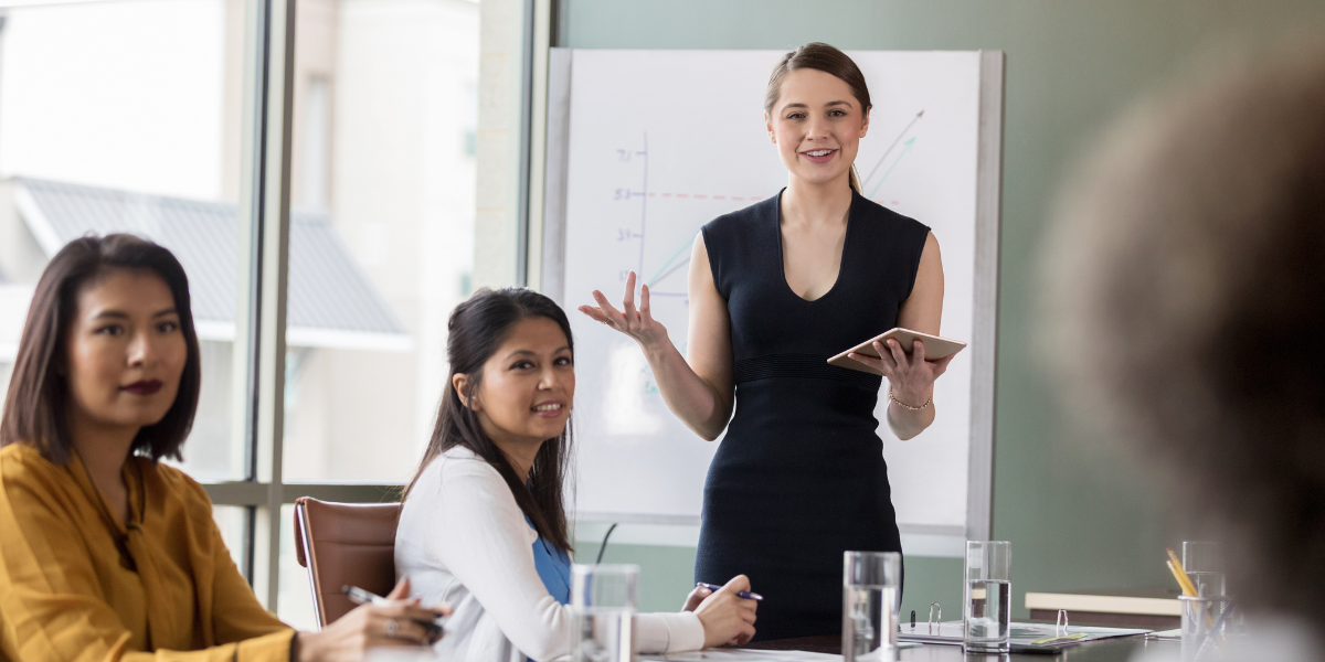 A woman standing holding a tablet and presenting to a boardroom of people.