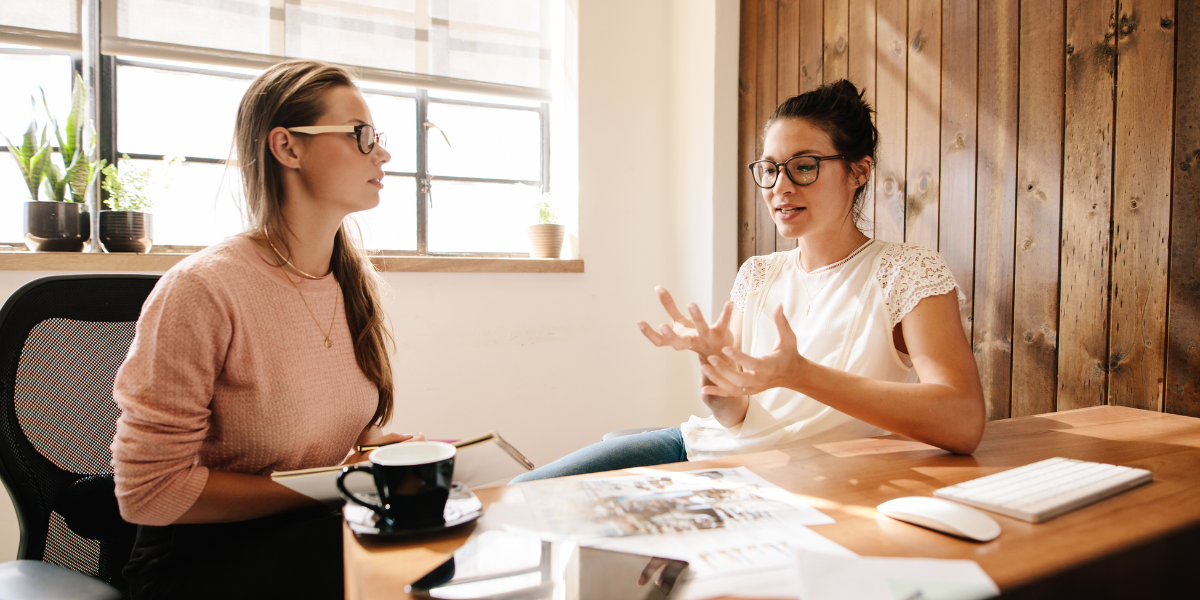 Two women are sat at a desk having a conversation and review paperwork.