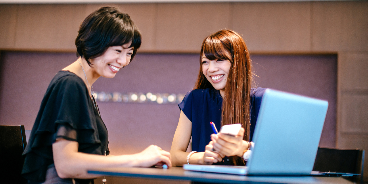 Two women are sat reviewing a laptop and conversing.