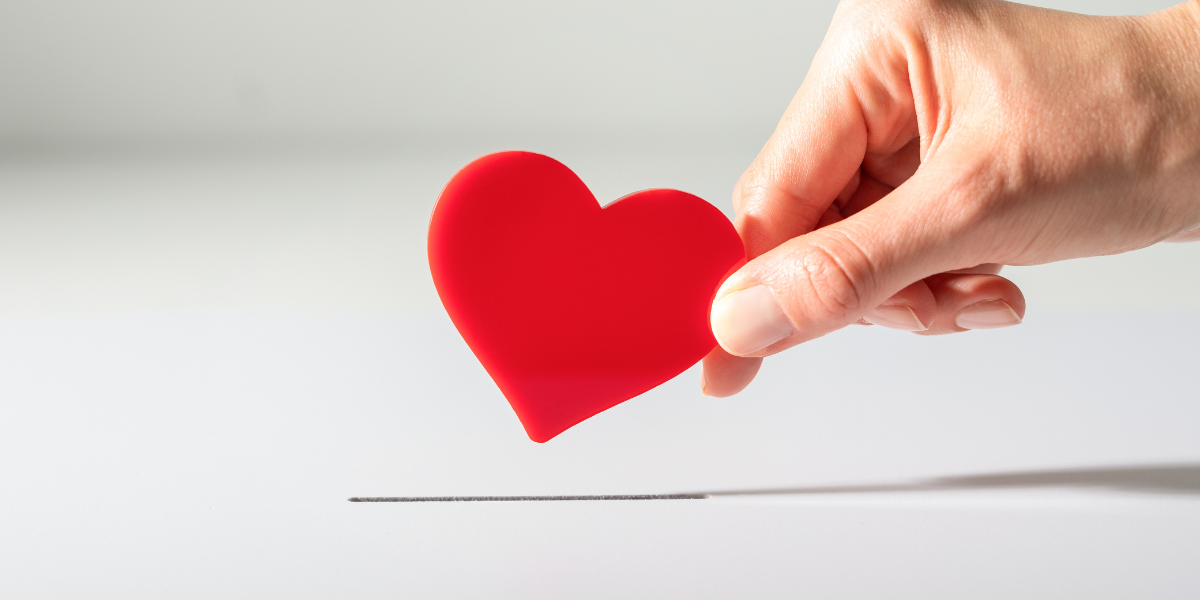 A hand is holding a red foam heart above a slot in a white box.