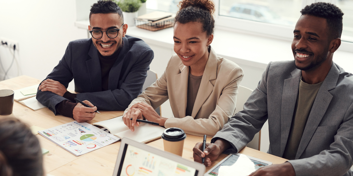 Three people are sat at a boardroom table working and conversing.