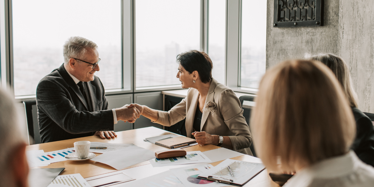 Two people are shaking hands in a boardroom during a meeting.