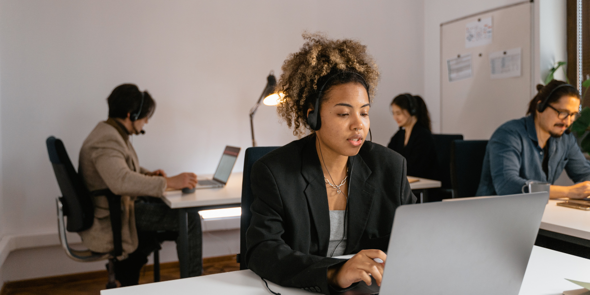 A woman sitting at a laptop working wearing headphones, while colleagues work in the background.