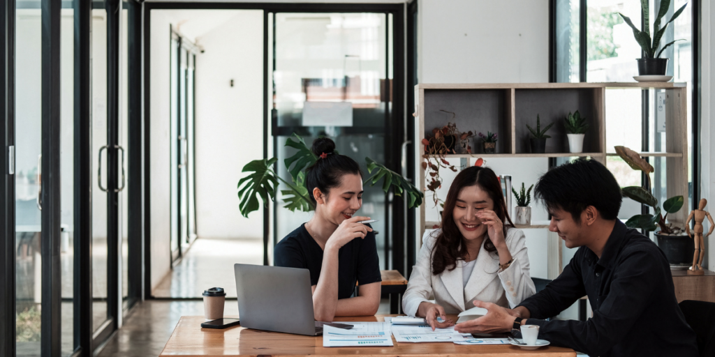 Three workers in an office space sitting at a boardroom table conversing.