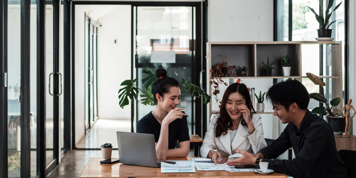 Three workers in an office space sitting at a boardroom table conversing.