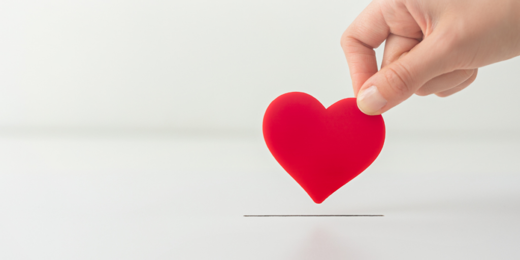 A hand is holding a red foam heart above a slot in a white box.