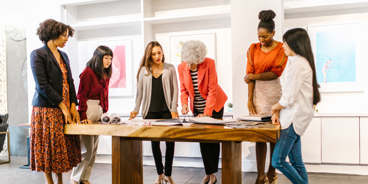 A group of women are standing at a boardroom table. They all appear to be collaborating.