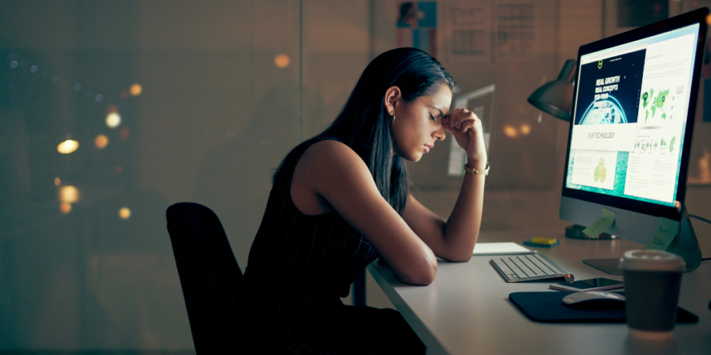 A woman is sat at her desktop computer it appears to be later in the night. The woman is resting her head on her hand.