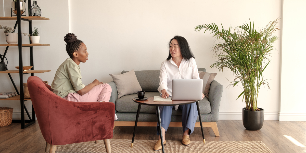 Two women are sat in an office conversing. One woman is sitting on a red velvet chair and the other is sitting on a grey couch.