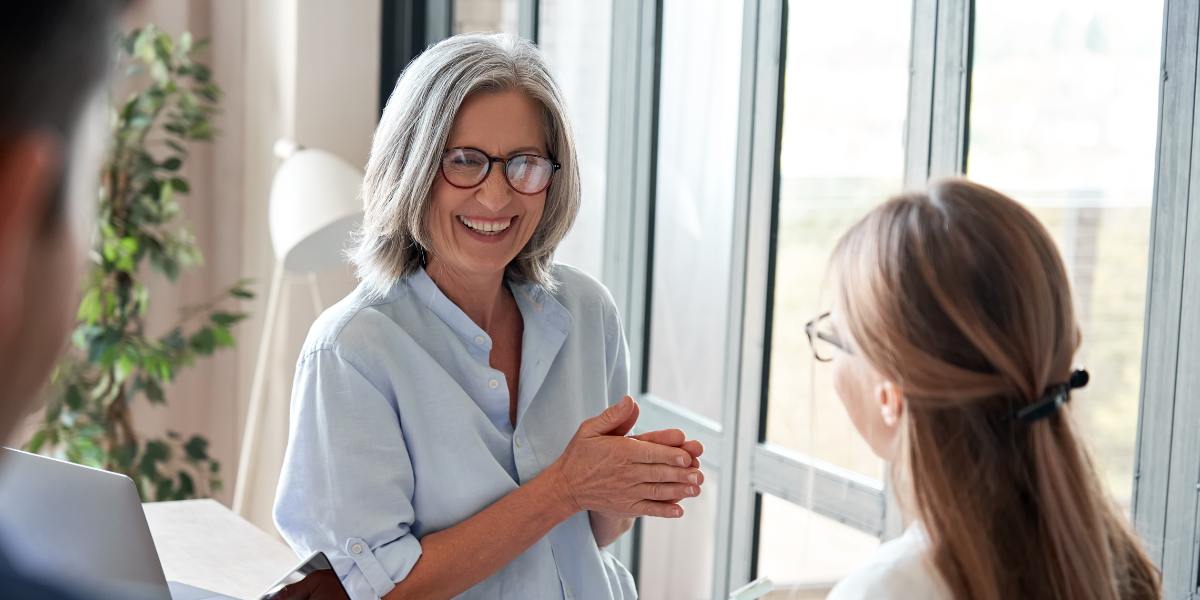 An older woman is sat at a desk facing another woman conversing.