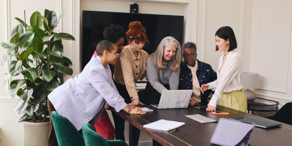 A group of women are standing in an office looking at a laptop on a desk conversing.