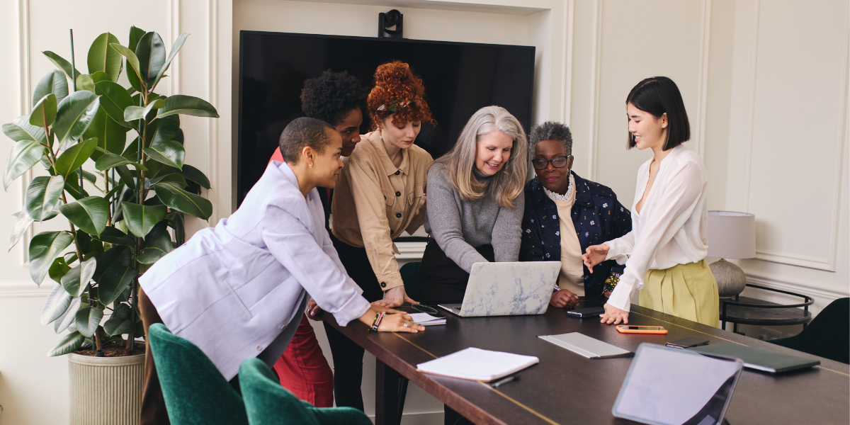 A group of women are standing in an office looking at a laptop on a desk conversing.